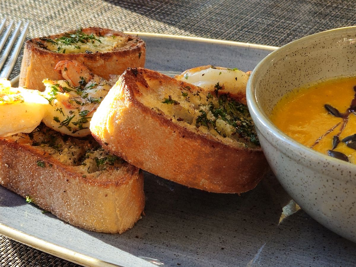 Geröstete Baguettescheiben mit Kräuterbutter und Garnelen auf einem Teller, daneben eine Schale mit Kürbissuppe