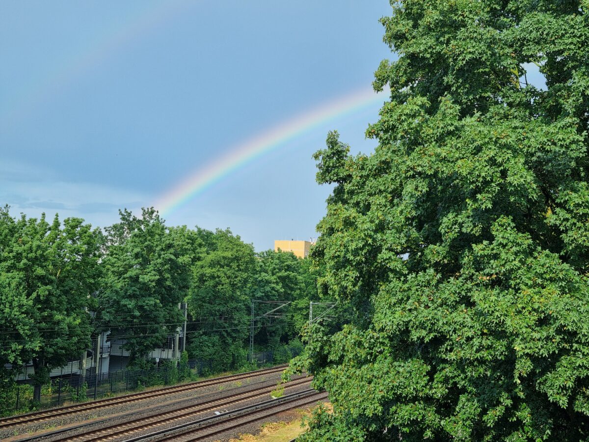 Regenbogen hinter Baumgrün und Bahngleisen